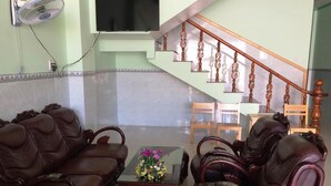 A living room with a brown leather sofa, a glass coffee table with a floral centerpiece, and a wooden chair. A TV is mounted on the wall, and a wooden staircase with a railing is visible in the background. - Y Thu Guesthouse (Phu Quoc)