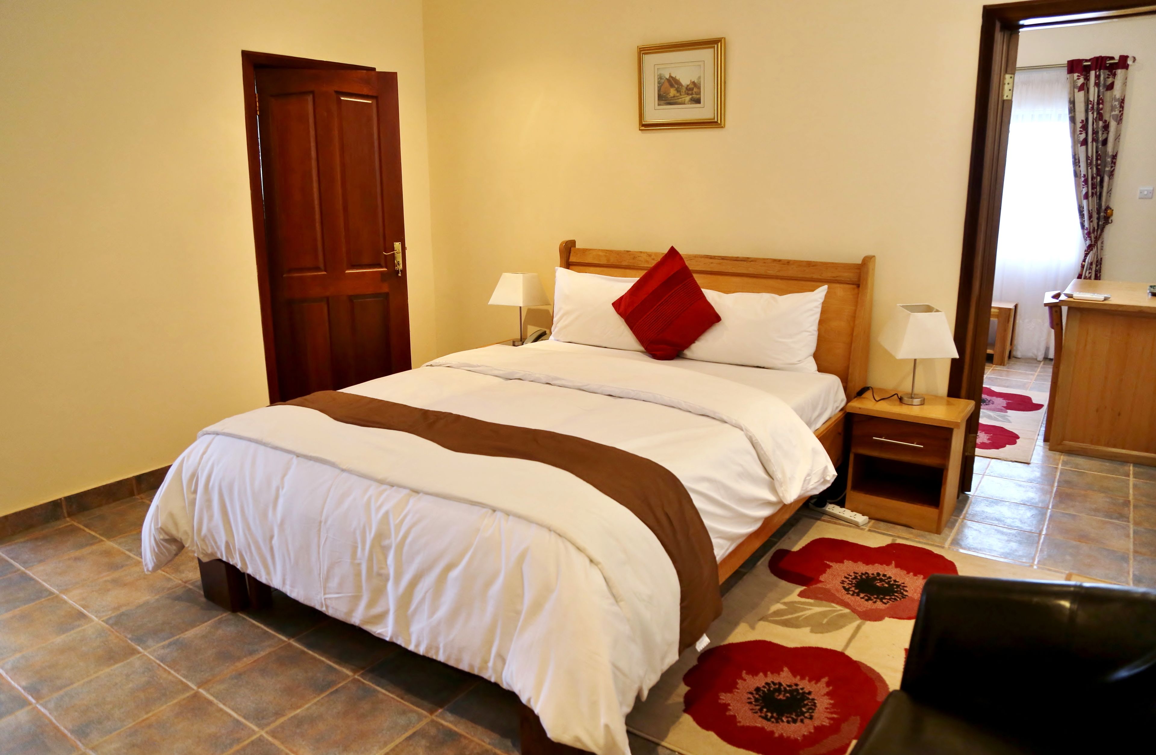 A neatly made bed with a wooden headboard, two bedside tables with lamps, a red pillow, and a floral rug on the floor.