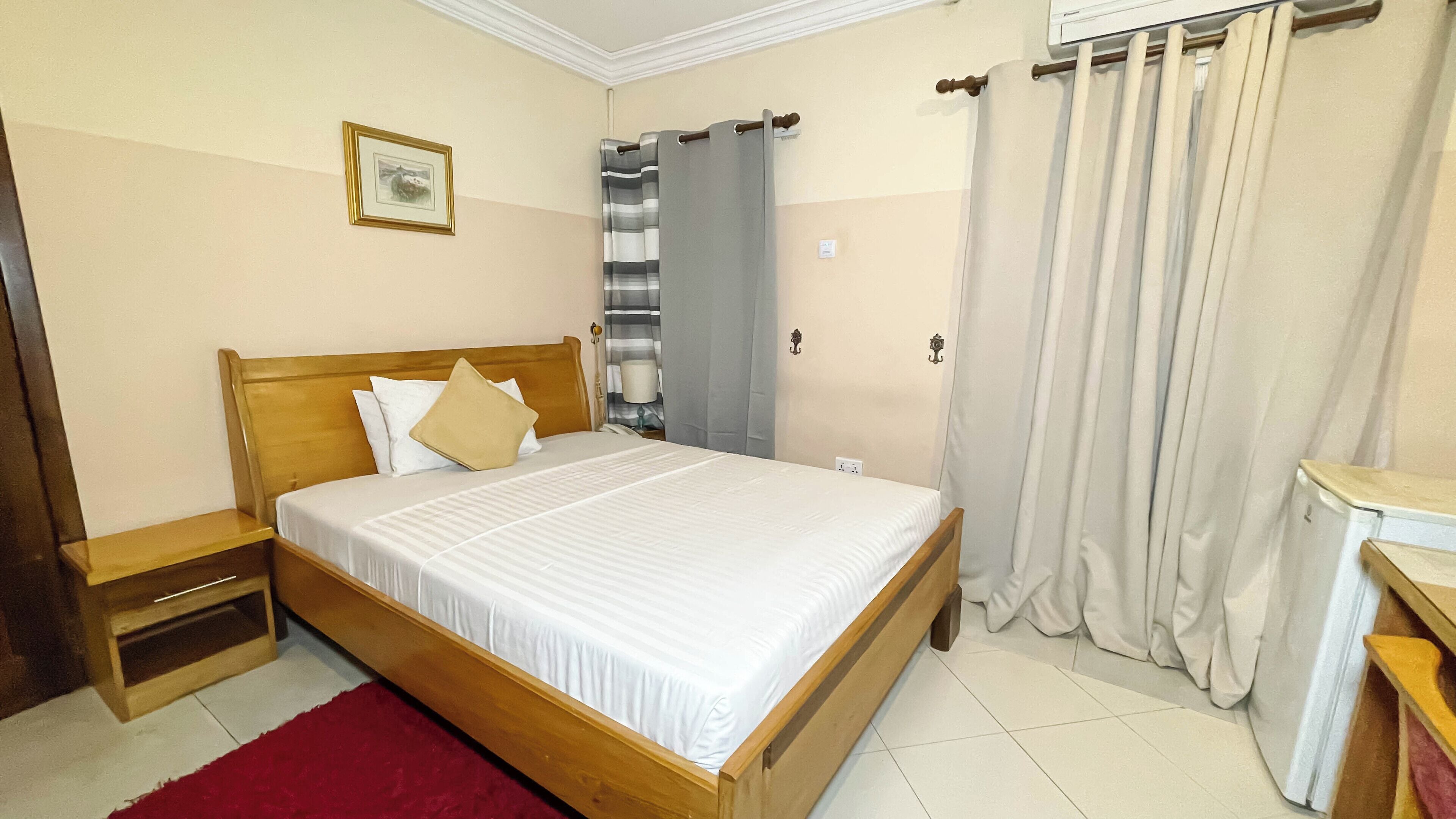A neatly made bed with white linens, a wooden headboard, and a red rug. There is a nightstand with a lamp, a framed picture on the wall, and a window with curtains.