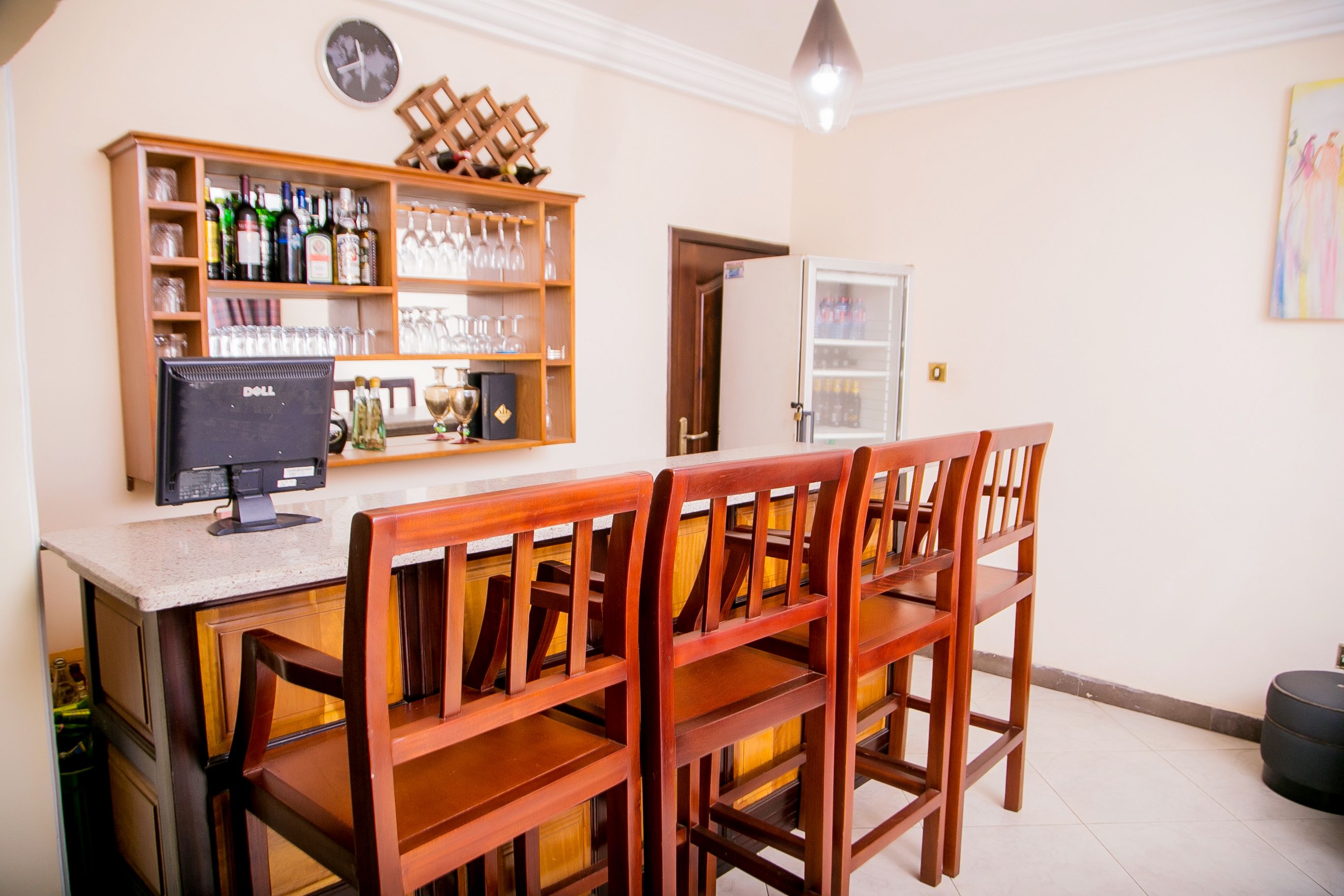 A bar area with wooden chairs and a counter, shelves with bottles and glasses, and a refrigerator.