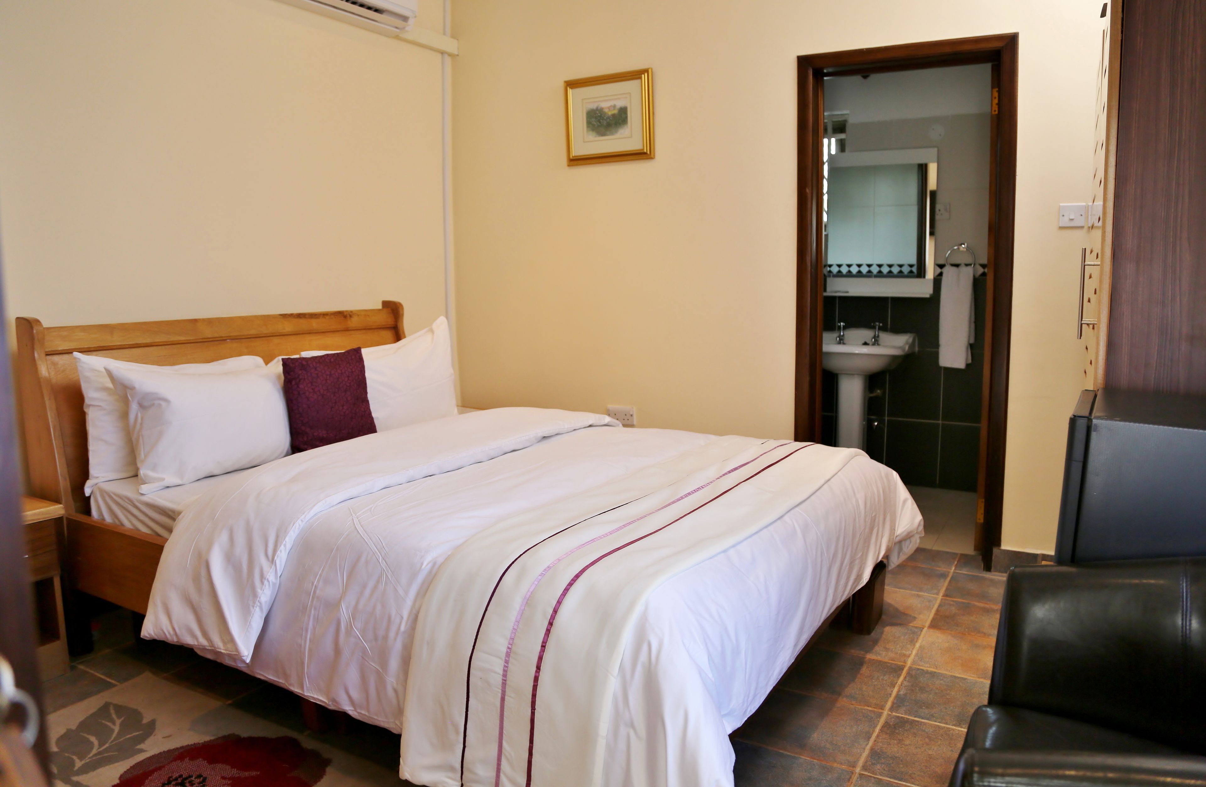 A neatly made bed with white bedding and a single red pillow, a wooden headboard, a small framed picture on the wall, and a bathroom visible through an open door.