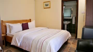 Una cama bien hecha con ropa blanca y una única almohada roja, un cabecero de madera, un pequeño cuadro en la pared y un baño visible a través de una puerta abierta.