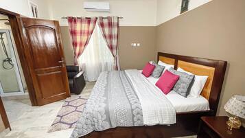 Un dormitorio con cama, un armario de madera, una silla, una mesita con lámpara y una ventana con cortinas.