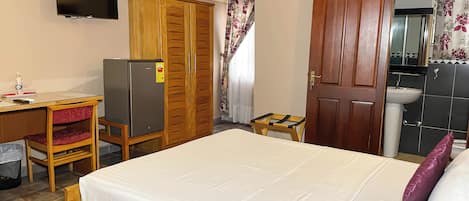 A bedroom with a bed, a wooden wardrobe, a desk with a chair, a small table, a television, and a bathroom with a sink and mirror.