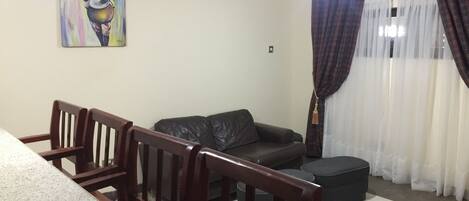 A living room with a dark leather sofa, wooden chairs, a painting on the wall, and a window with curtains.