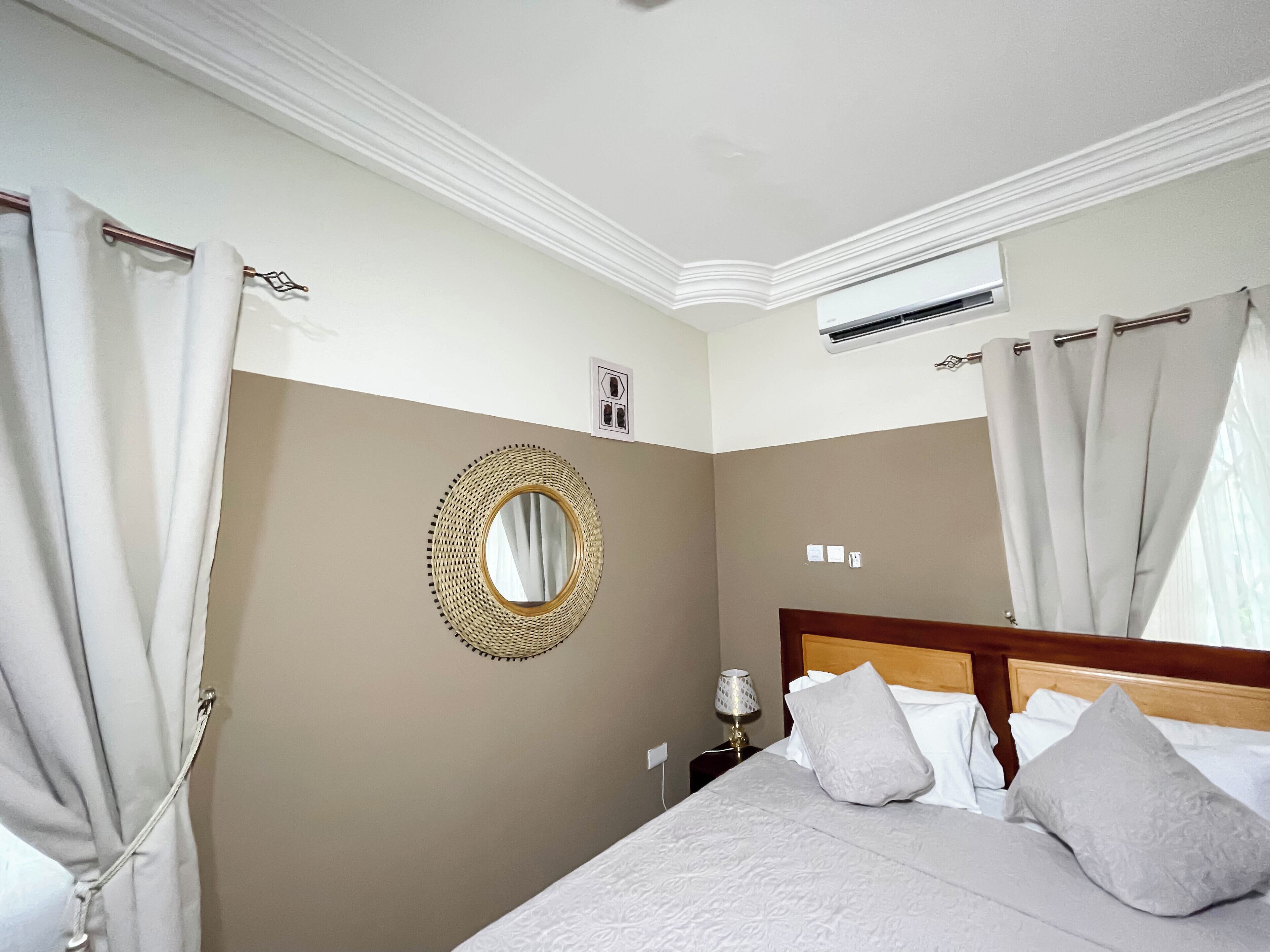 Quarto com cama, cortinas, espelho redondo, ar condicionado e mesa de cabeceira com abajur.