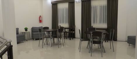 Un comedor moderno y bien iluminado con suelo de baldosas limpias, un sofá, una mesita con una planta y un extintor en la pared.