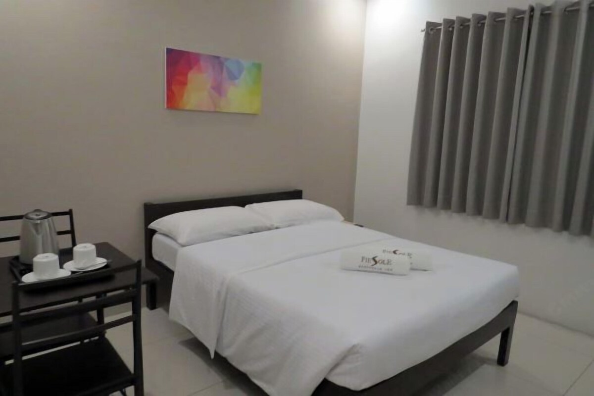 Une chambre d’hôtel avec un lit, une table de chevet sur laquelle se trouvent une bouilloire, des tasses et un plateau, ainsi qu’une peinture abstraite aux couleurs vives accrochée au mur.