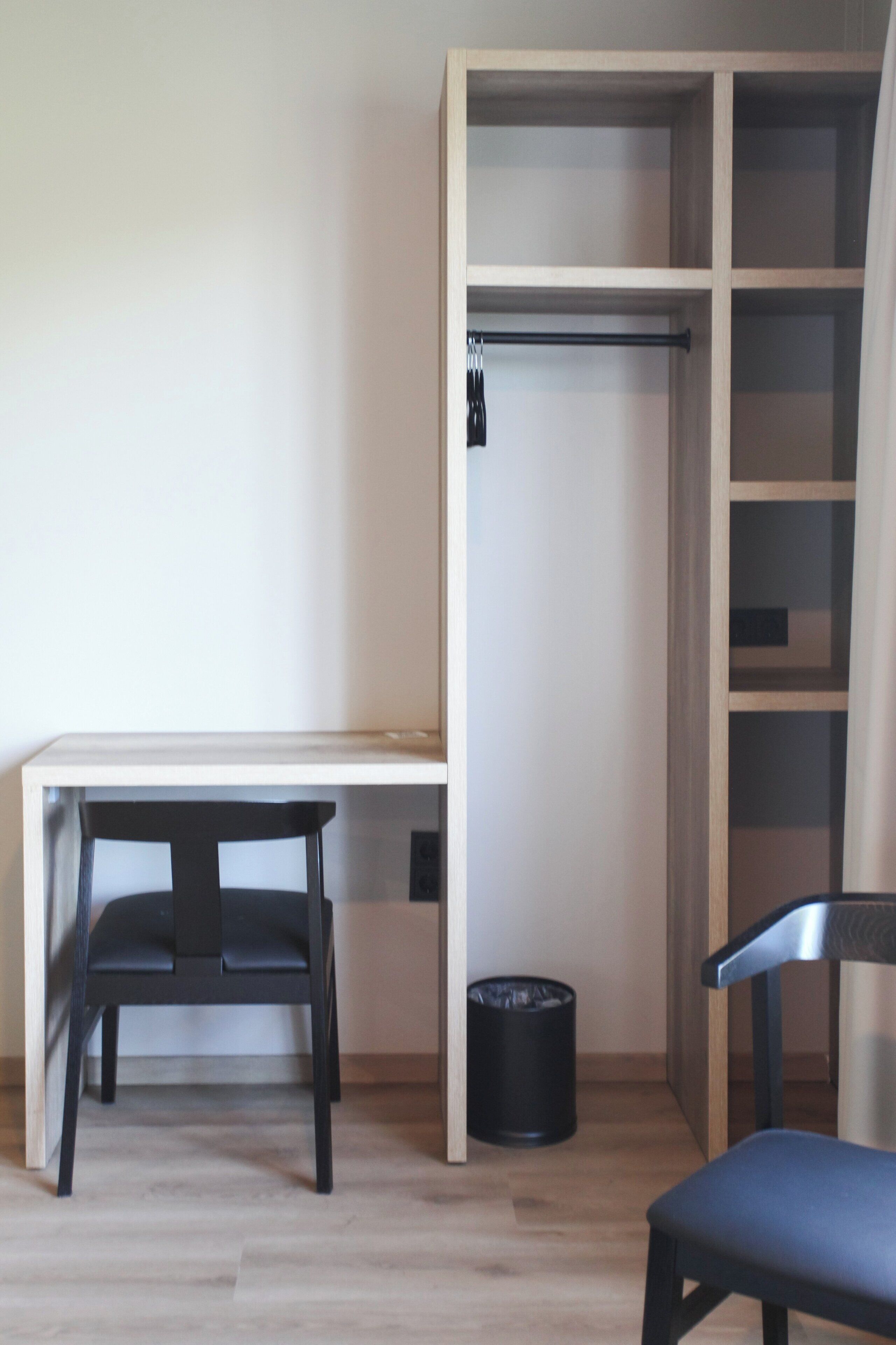 A room with a desk, two chairs, a black trash bin, and a wooden wardrobe.