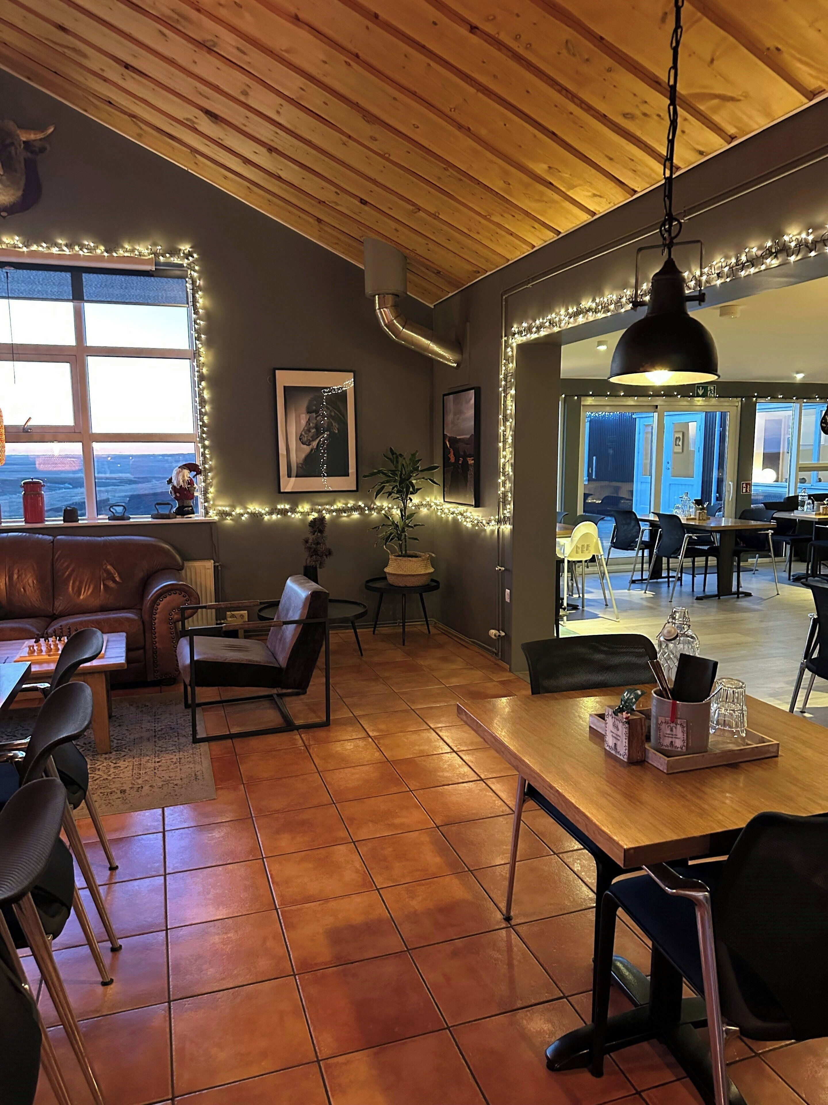 A cozy living area with a fireplace, a dining table, and a sofa. There is a large window with a view, and the room has wooden beams on the ceiling.