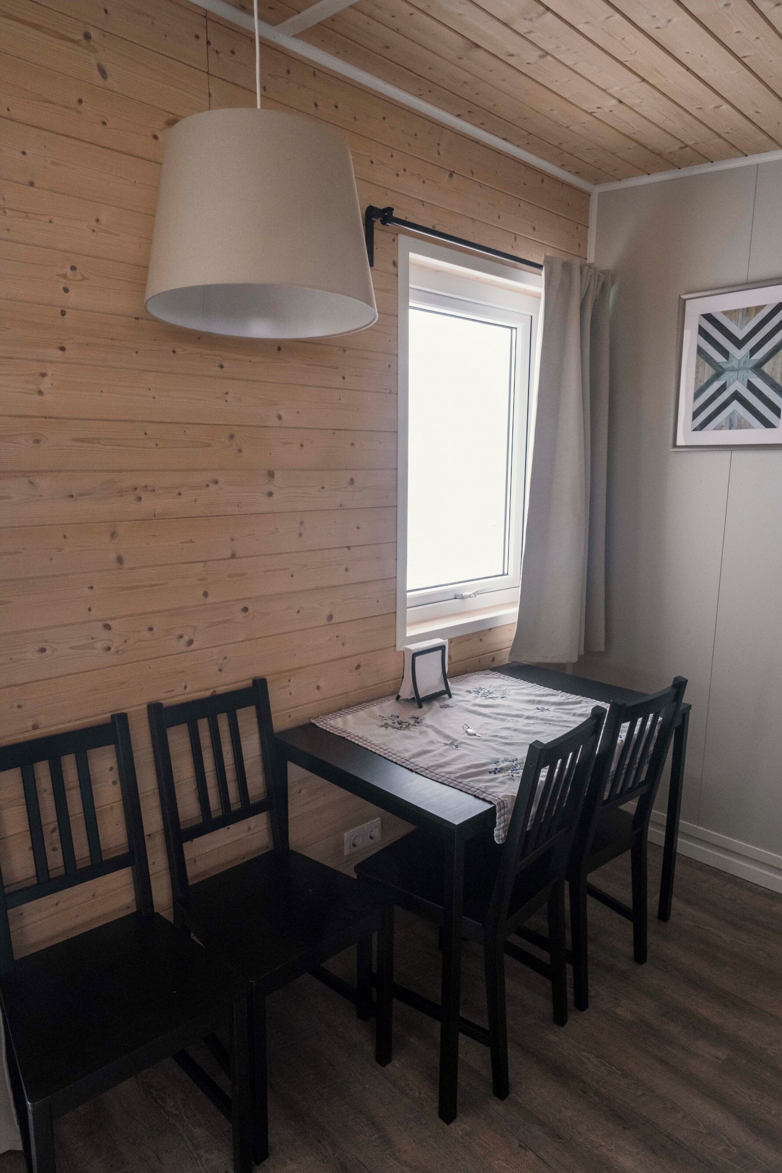 Un comedor con una mesa de madera, cuatro sillas, una lámpara colgante, una ventana con cortinas y un cuadro en la pared.
