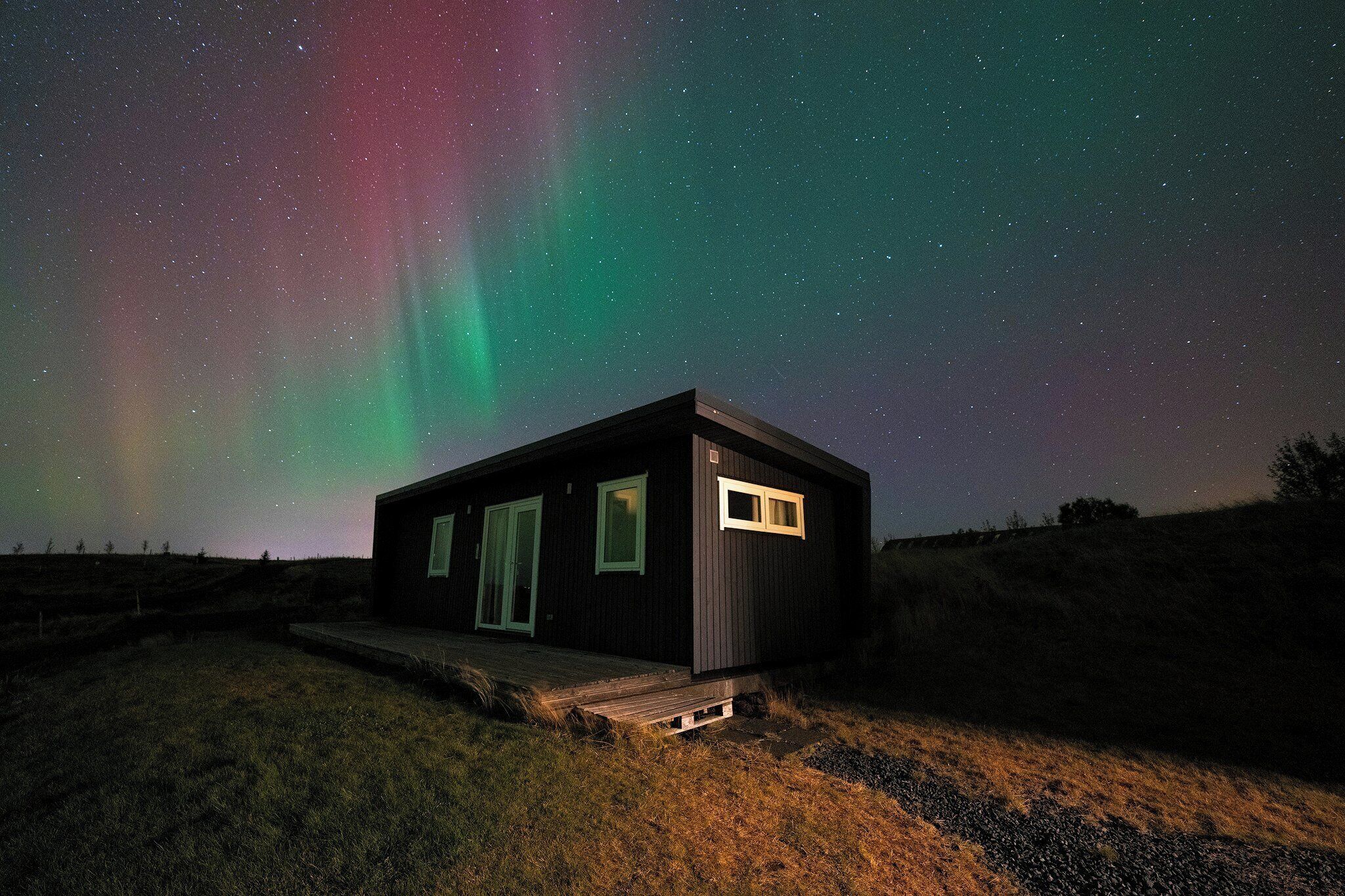 Eine kleine Holzhütte mit einem Fenster und einer Tür, umgeben von einer Wiese unter einem sternenübersäten Nachthimmel mit Nordlichtern.