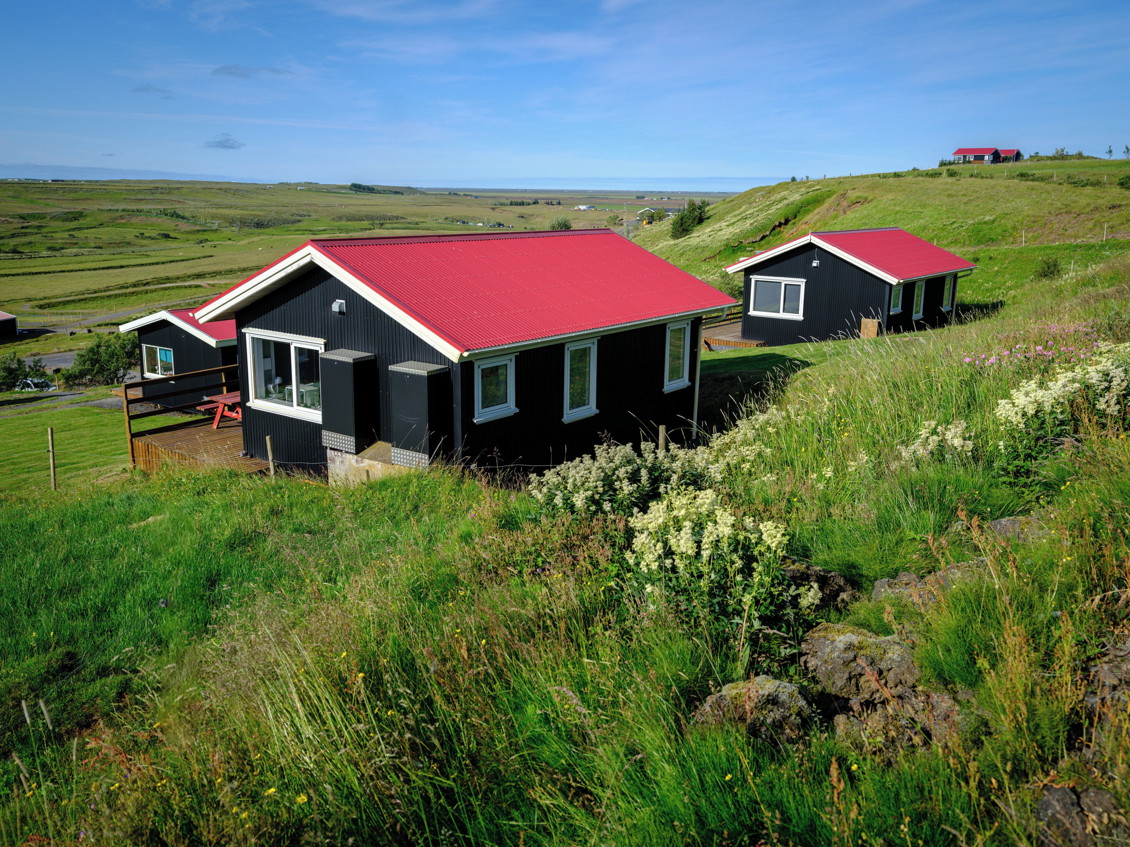 A black house with a red roof and a wooden deck, surrounded by green fields and hills.
