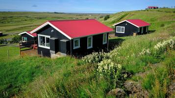 A black house with a red roof and a wooden deck, surrounded by green fields and hills.