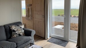 Ein Zimmer mit grauem Sofa, einer Holztür und Blick auf ein grünes Feld durch eine Glastür.