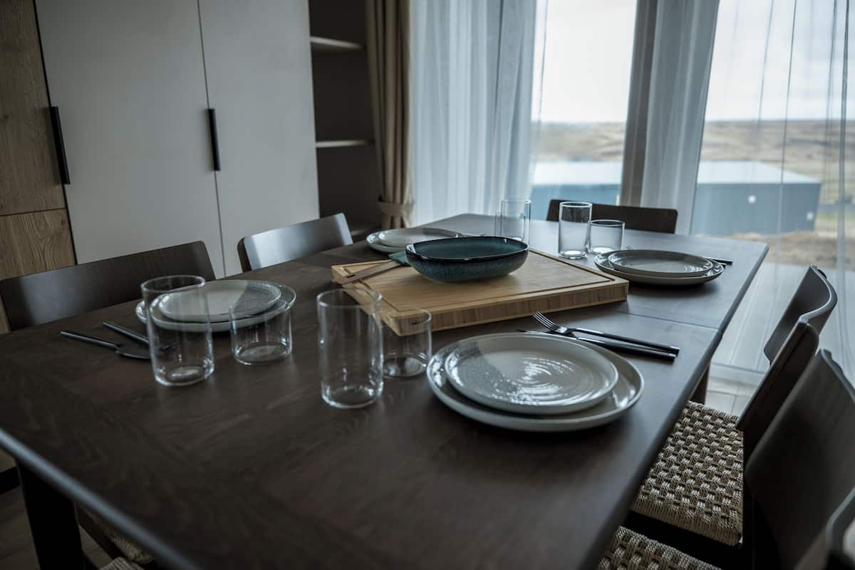 Mesa de jantar posta com pratos, copos e uma tigela, com vista para o exterior através de grandes janelas.