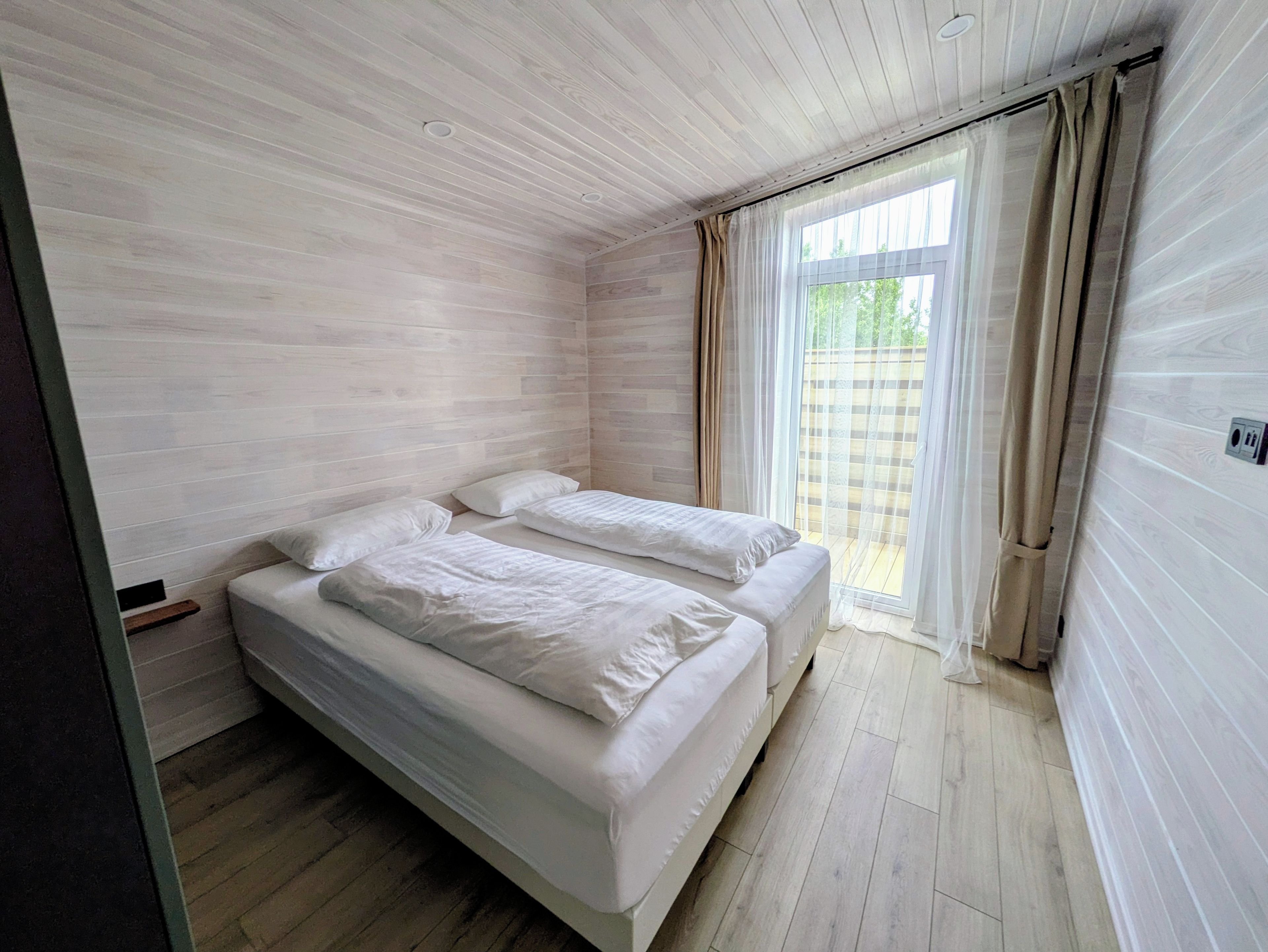 A room with a bed, a window with curtains, and a view of greenery outside.