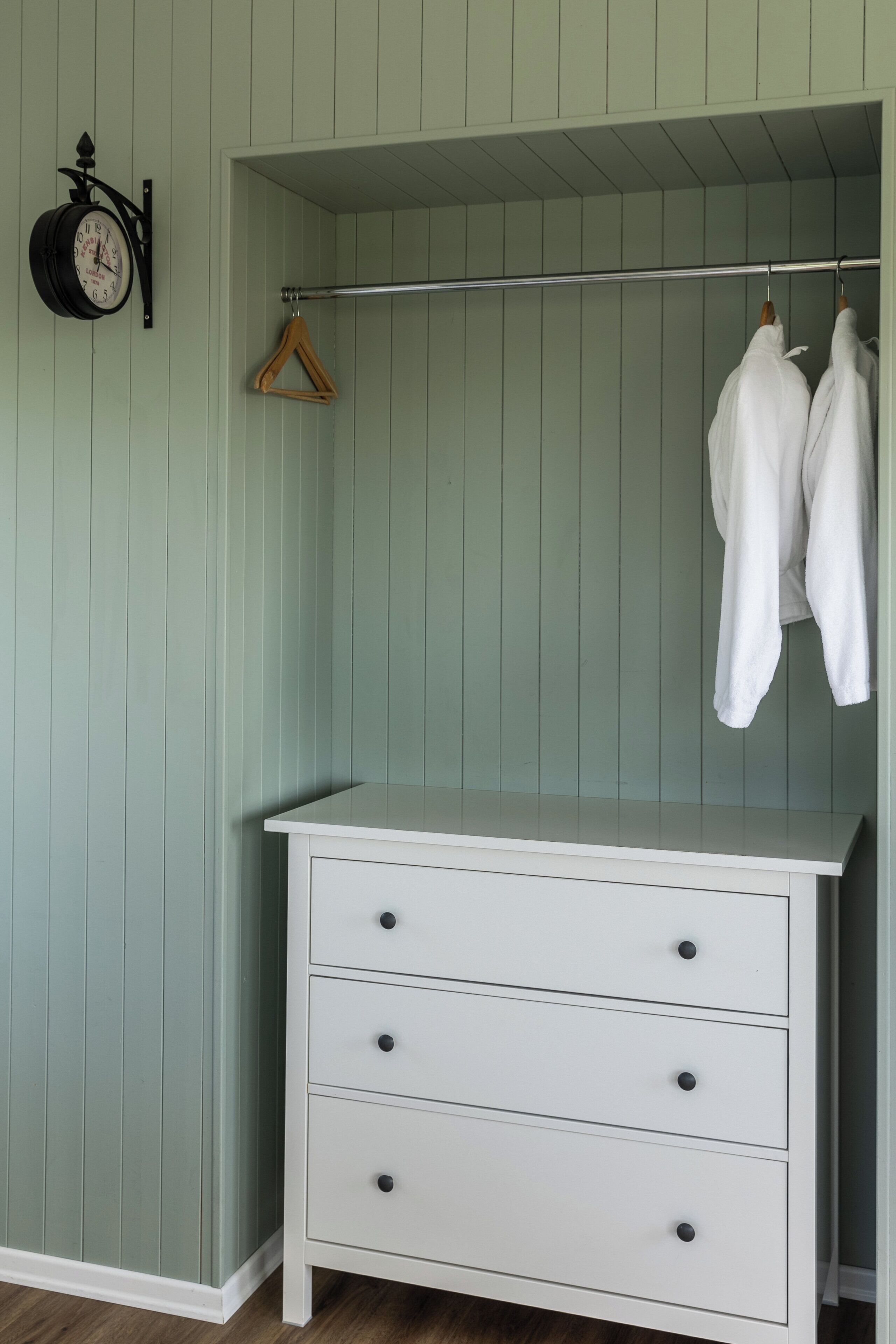 A white dresser with drawers, a coat rack with hangers, and a wall-mounted clock in a room with vertical paneling.