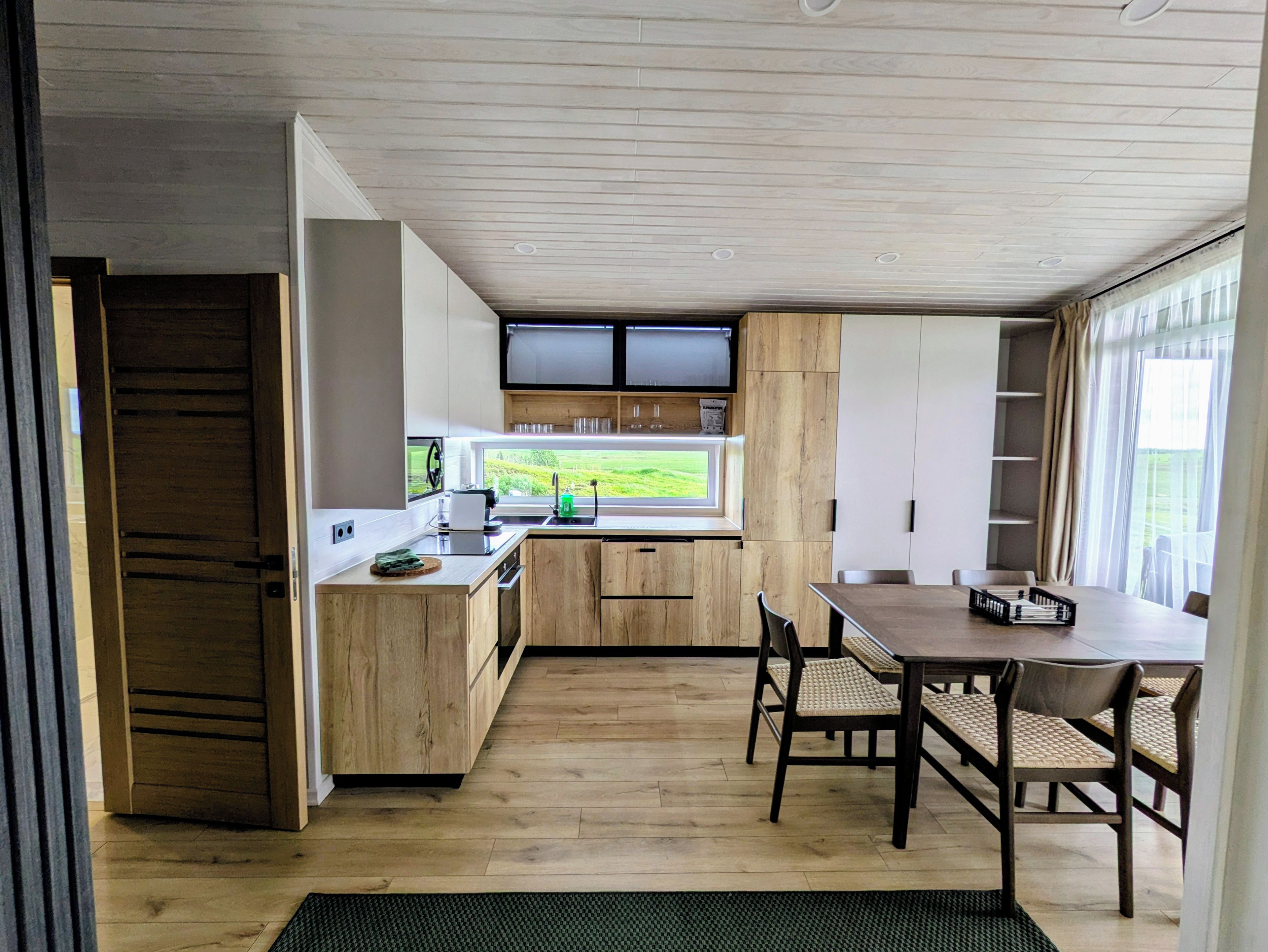 A modern kitchen with wooden cabinets, a dining area with a table and chairs, and a view of greenery outside.