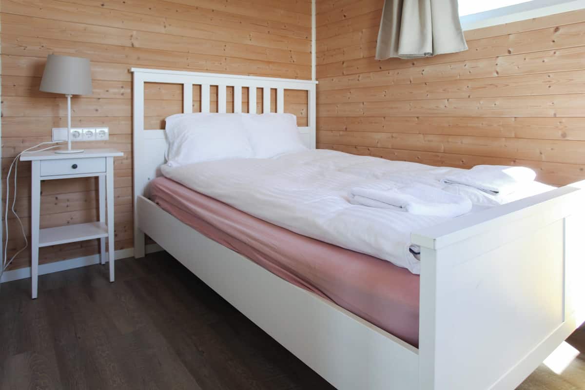 Quarto com parede de madeira, cama branca, mesa de cabeceira com abajur e tomada.