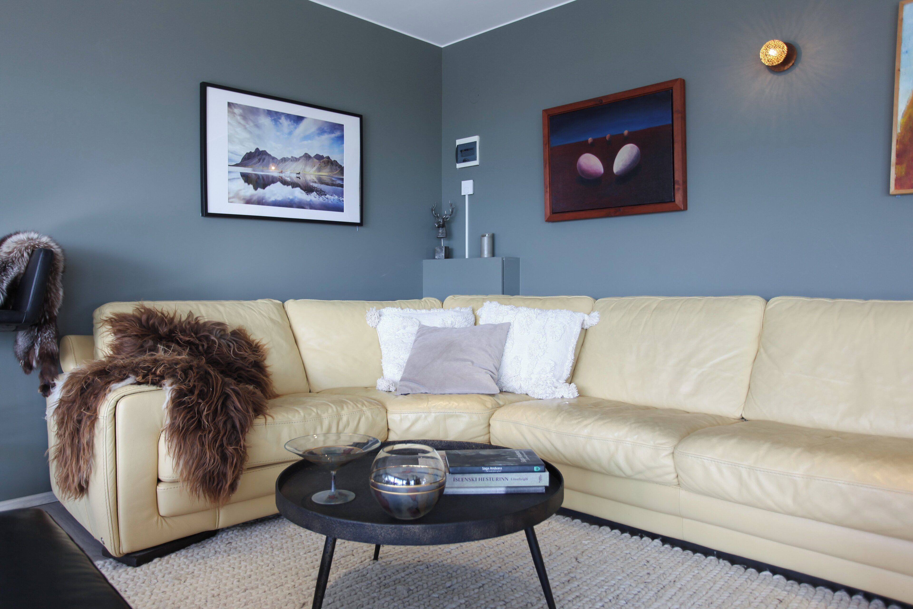 Een woonkamer met een beige bank, een bijzettafeltje met boeken en glazen, en ingelijste foto's aan de muur.