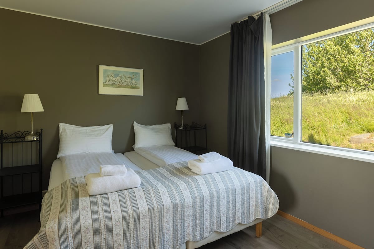 Uma cama de casal com colcha listrada, duas mesas de cabeceira com abajures, um quadro na parede e uma janela com vista para o verde.