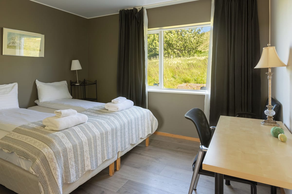 Quarto de hotel com cama, escrivaninha, cadeira, luminária e janela com cortinas.