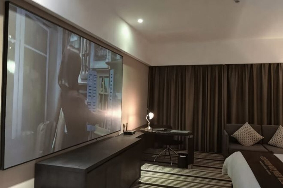 A modern hotel room with a large flat-screen TV, a sofa, a desk with a lamp, and a bed with a striped rug.