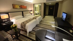 Quarto de hotel com duas camas, uma televisão, uma escrivaninha e um banheiro visível através de uma porta de vidro.