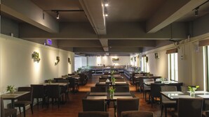 Um restaurante amplo com várias mesas e cadeiras, uma área de bar ao fundo e plantas decorativas nas paredes.