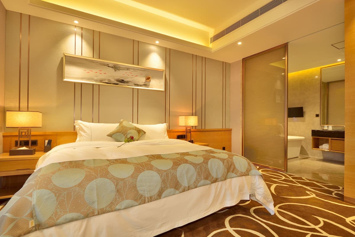 A hotel room with a large bed, bedside lamps, a nightstand, a mirror, and a bathroom visible through a sliding door.