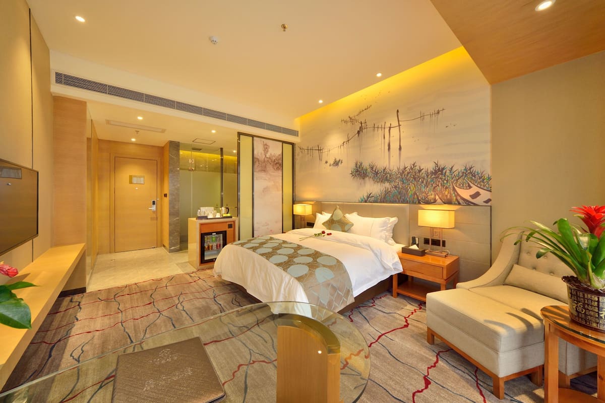 A modern hotel room with a large bed, a sitting area, a desk, and a bathroom.