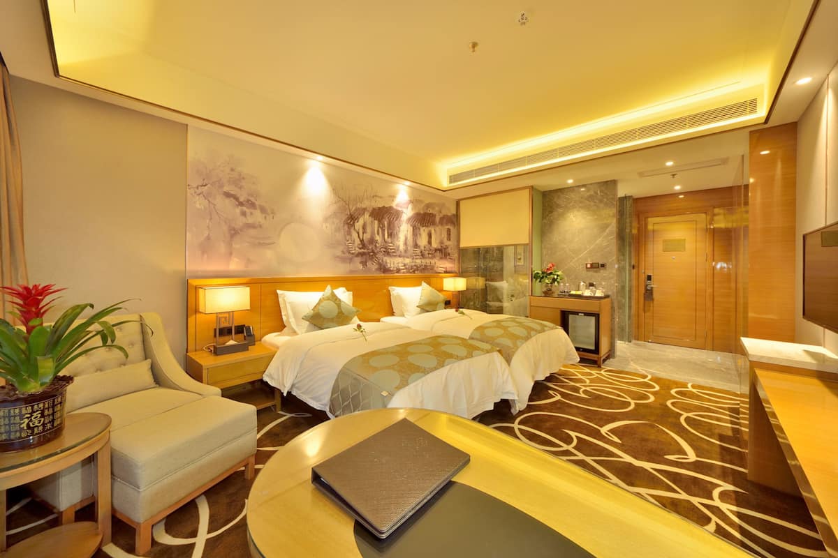 A hotel room with a large bed, two bedside tables with lamps, a desk with a laptop, a potted plant, and a large painting on the wall.