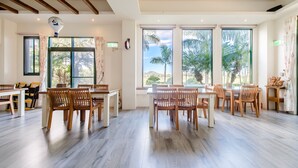 A spacious dining area with wooden tables and chairs, large windows with views of palm trees, and a ceiling fan.