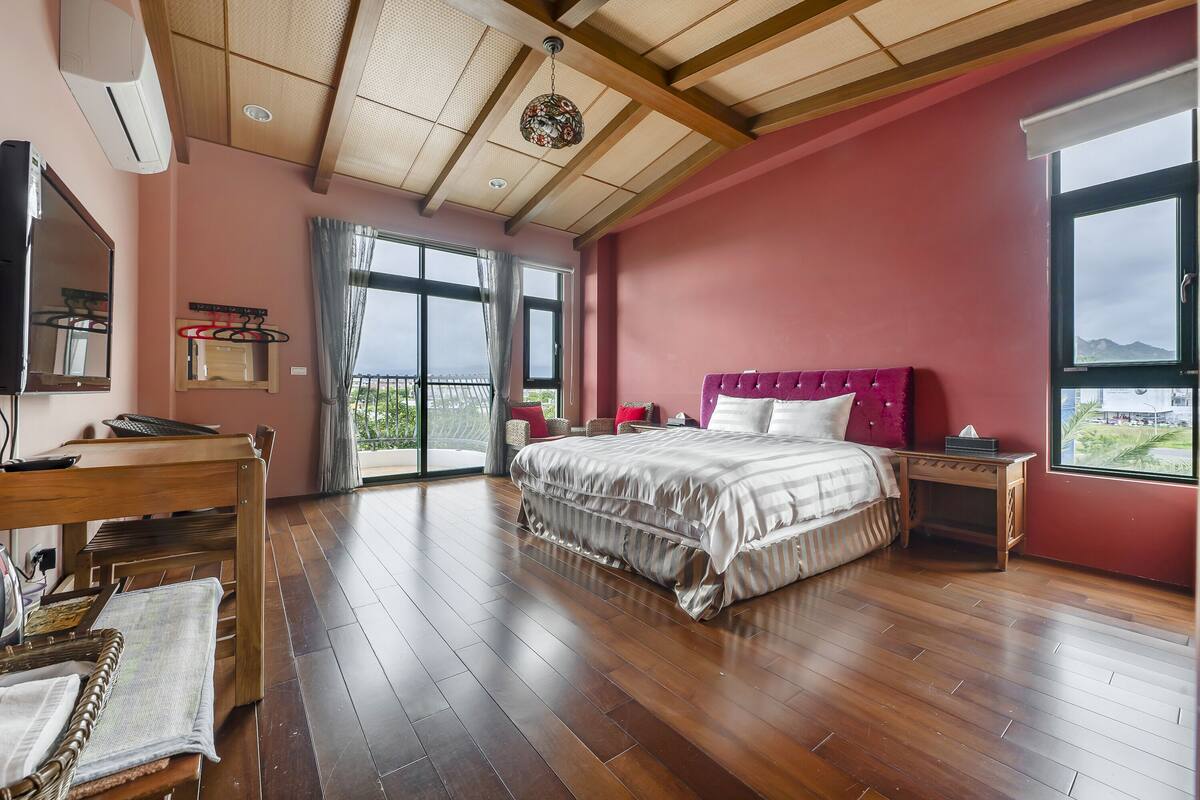 A bedroom with a large bed, a wooden desk, a chair, a TV, and a window with a view.