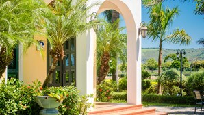 A building with a covered entrance, steps leading up to it, and a view of a landscaped garden with palm trees and a distant hill.