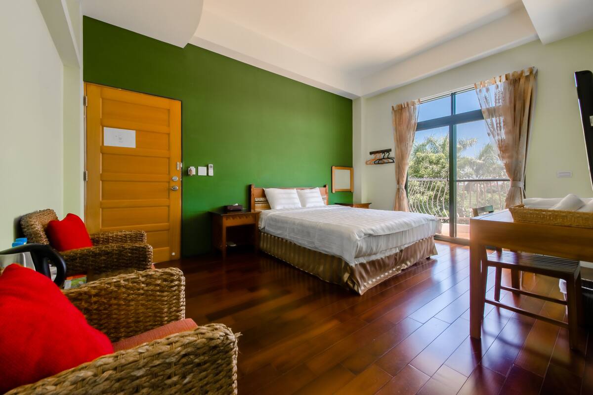 A room with a bed, wicker chairs, a wooden table, and a large window with curtains.