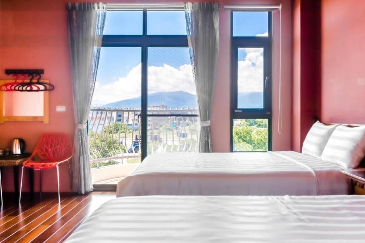 A hotel room with a large bed, a red chair, a table with a kettle, and a balcony with a view of a mountain and cityscape.
