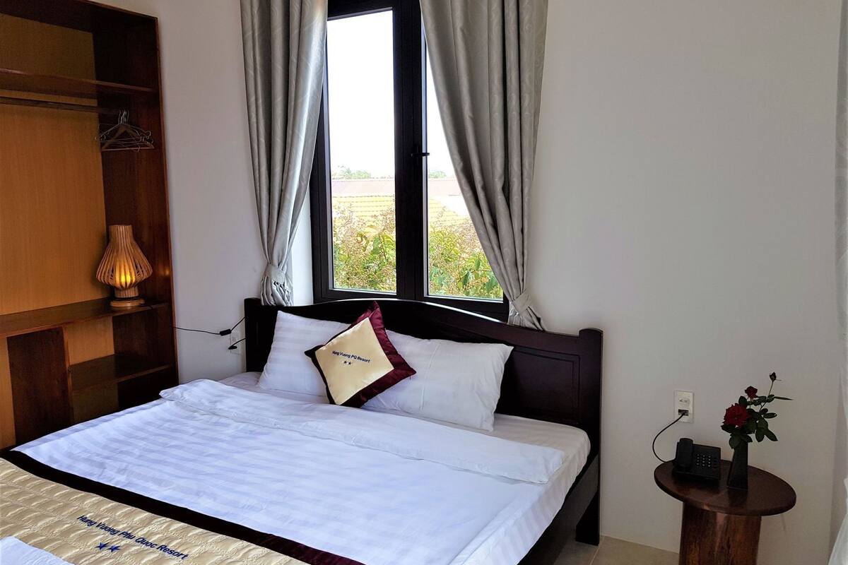 A hotel room with a bed, a bedside table with a phone and a vase, a lamp, and a window with curtains.
