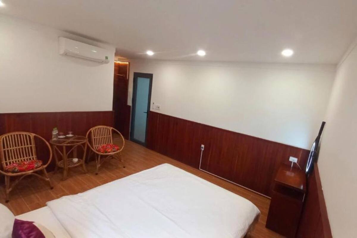 A hotel room with a bed, two wicker chairs, a small table, a TV, and an air conditioning unit.