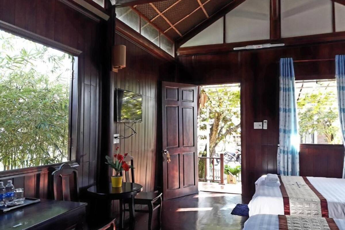 A room with a wooden ceiling, a bed with a red and white bedspread, a small table with a vase of flowers, and a view of greenery outside.
