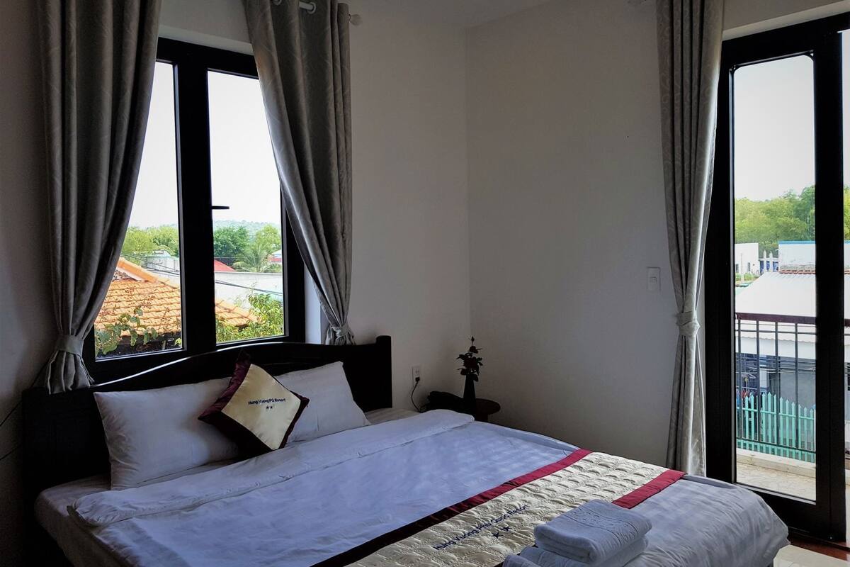 A hotel room with a bed, curtains, a nightstand, a lamp, and a view of buildings and greenery outside.