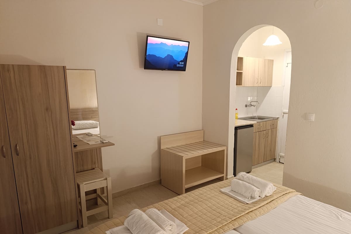 Une chambre d’hôtel comprenant un lit, un bureau, une chaise, une télévision, une petite table et un coin cuisine visible à travers une arche.