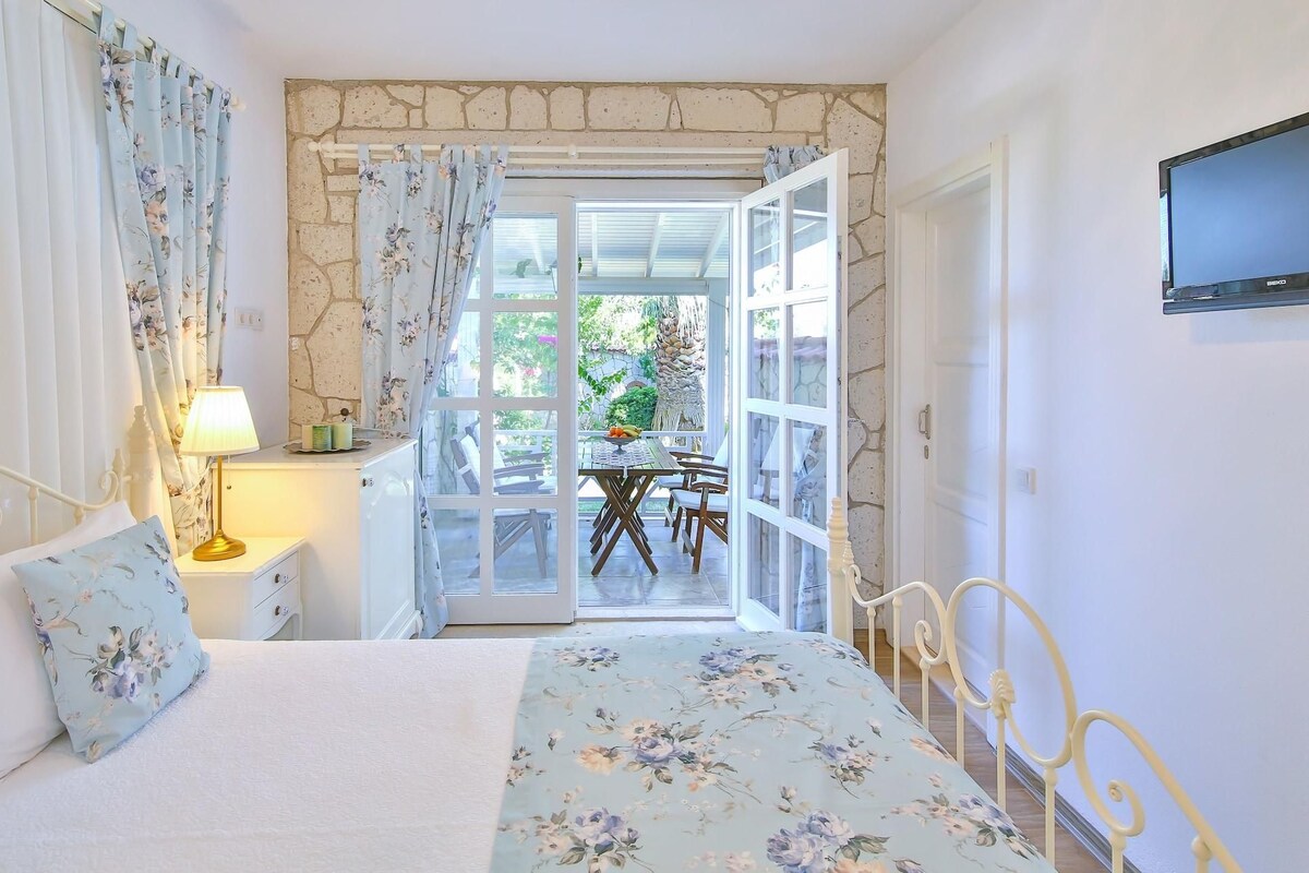 A bedroom with a bed, a nightstand, a lamp, a TV, and a view of an outdoor patio with a table and chairs.