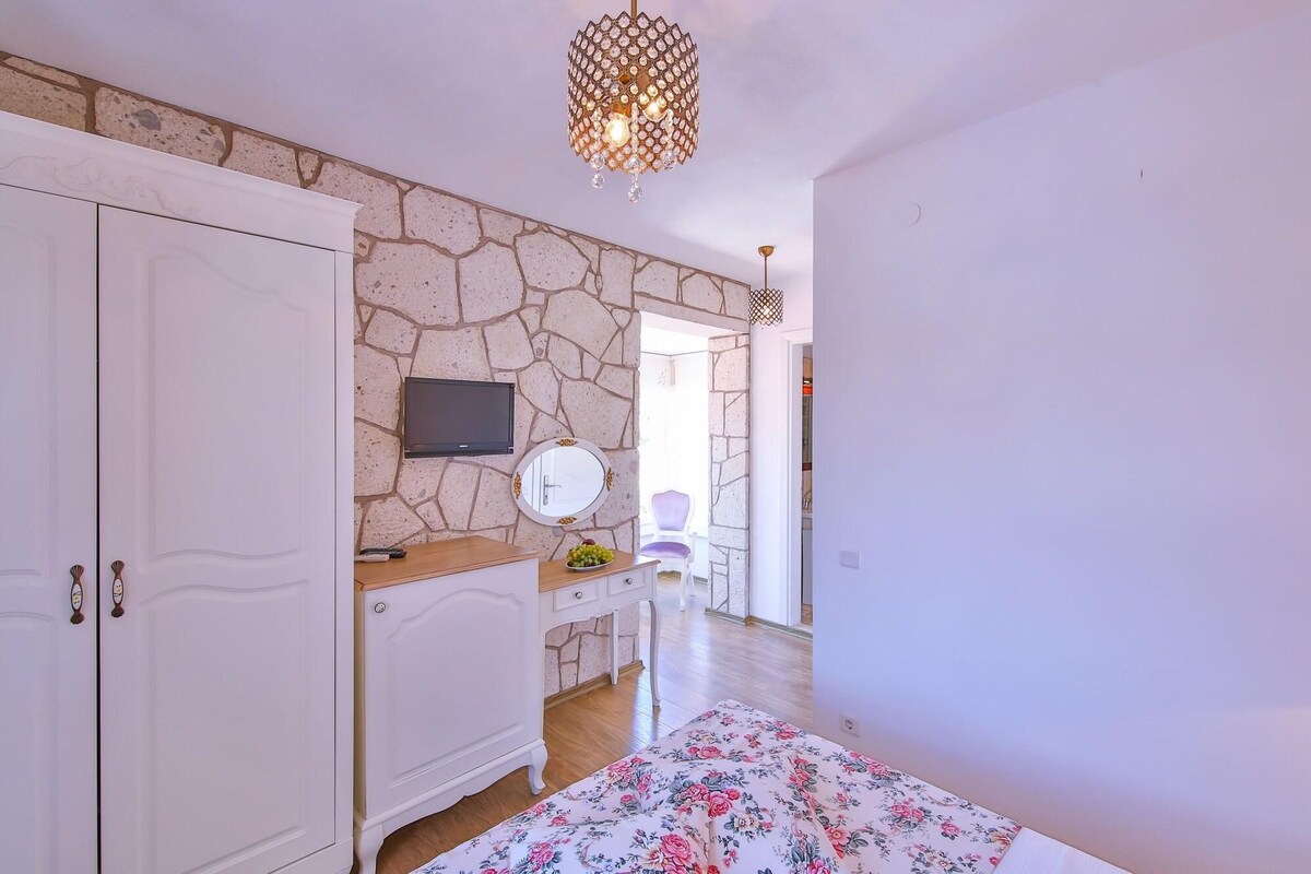 A bedroom with a stone wall, a TV, a mirror, a bed with a floral bedspread, and a wooden dresser.