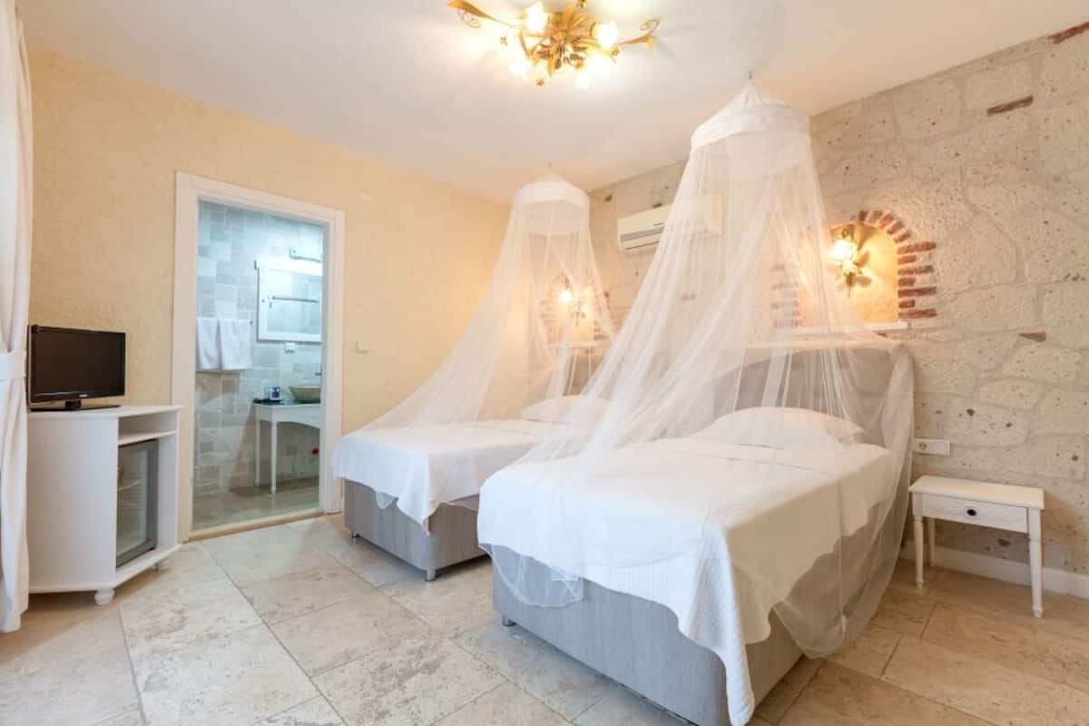 Quarto com duas camas, televisão, escrivaninha e banheiro visível através de uma porta de vidro.