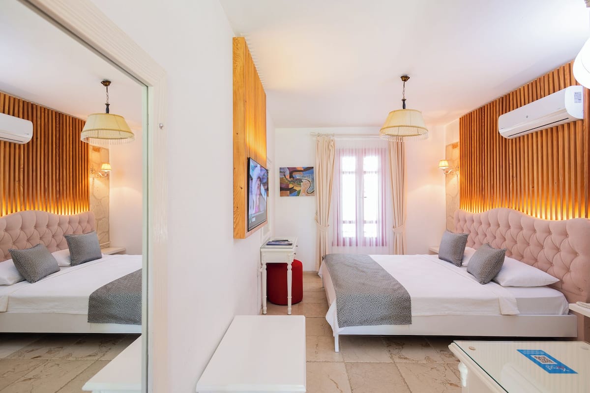 Quarto de hotel com duas camas, um espelho, uma mesinha e uma janela com cortinas.