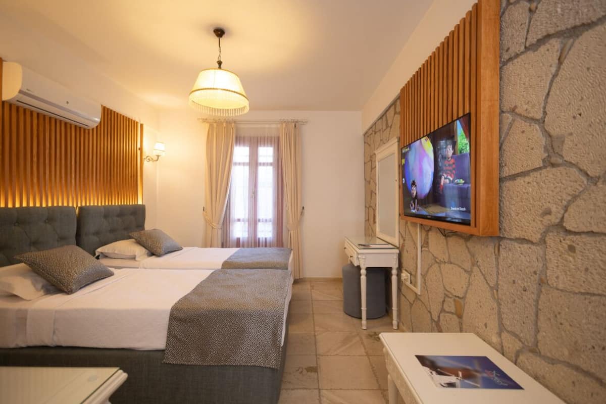 Quarto de hotel com duas camas, uma televisão presa a uma parede de pedra e uma mesinha de centro com uma revista.