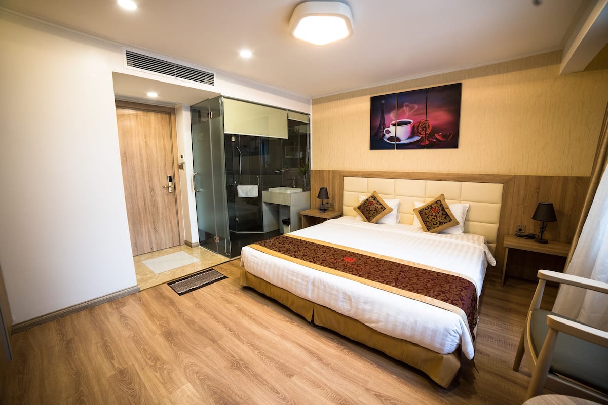 A hotel room with a large bed, a shower area, a bedside table with a lamp, and a painting on the wall.