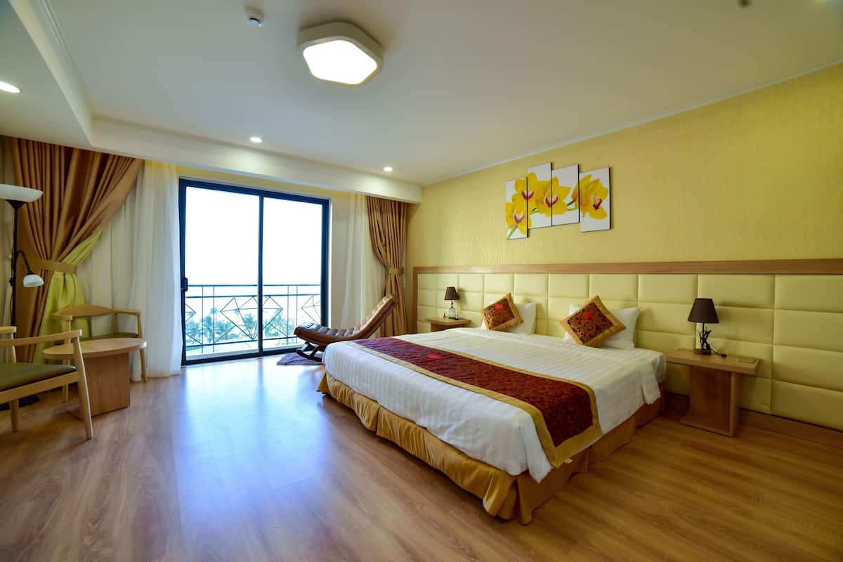 A spacious hotel room with a large bed, a sitting area with a chair, a small table, and a view of the outdoors through a large window.
